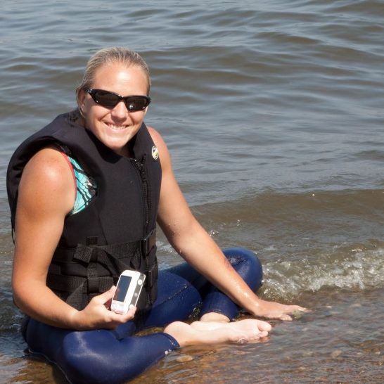 Adult female skier sitting at the edge of the lake; she has her camera and a HUGE smile on her face