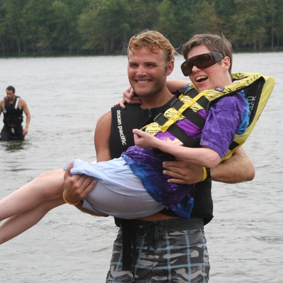 an adult male volunteer is carrying a skier to shore from the water; both have HUGE smiles on their faces