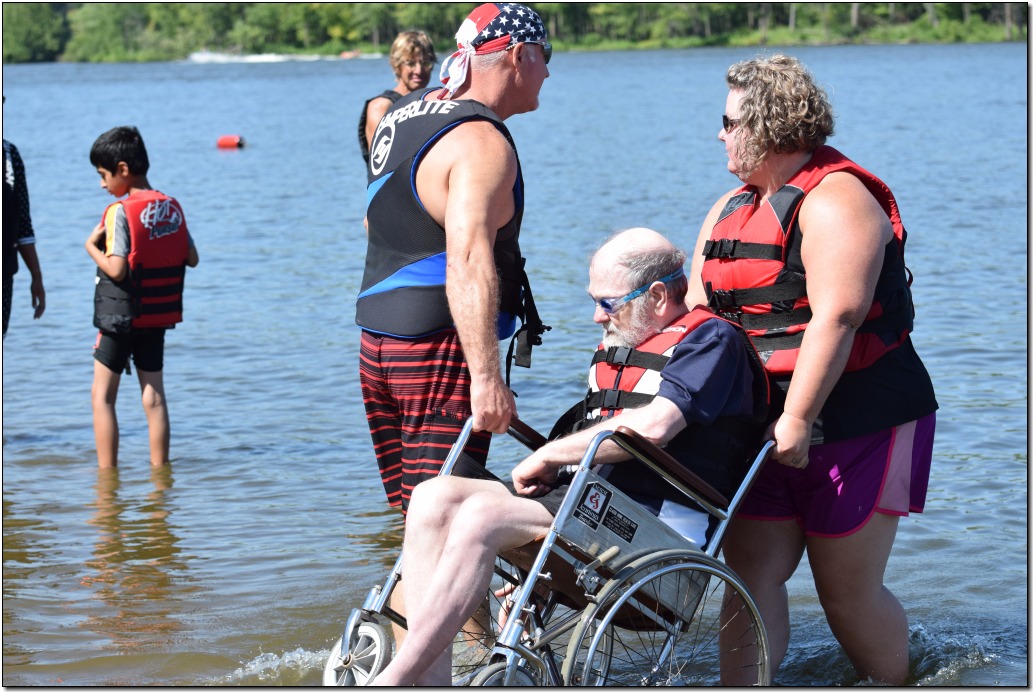 two adult volunteers transferring a male skier, in a wheelchair, from the beach to the water