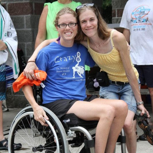 Two young females, one in a wheelchair, have their arms around each other and are smiling at the camera
