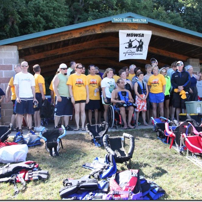 group photo of skiers and volunteers, under the pavillion with the MDWSA banner hanging from the rafters and all of the ski equipment and life jackets on the ground
